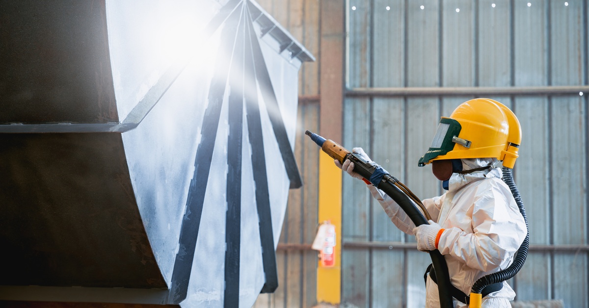 A person wearing a white protective suit and a yellow safety helmet holds a black tube that is ejecting a spray.