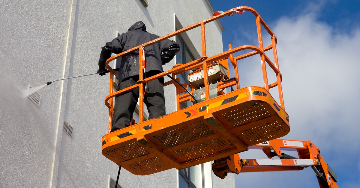 A person dressed in all black stands on an orange, motorized lift. They're using a hydroblaster on the side of a building.