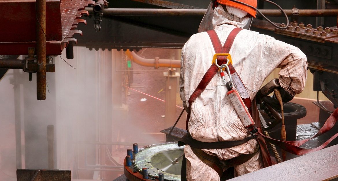 A person wearing a white protective suit, a red safety harness, and an orange safety helmet is hydroblasting a pipe.