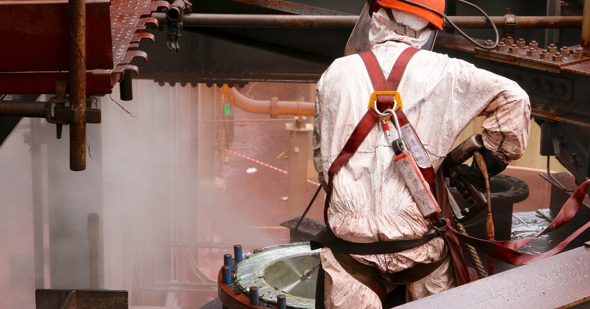 A person wearing a white protective suit, a red safety harness, and an orange safety helmet is hydroblasting a pipe.