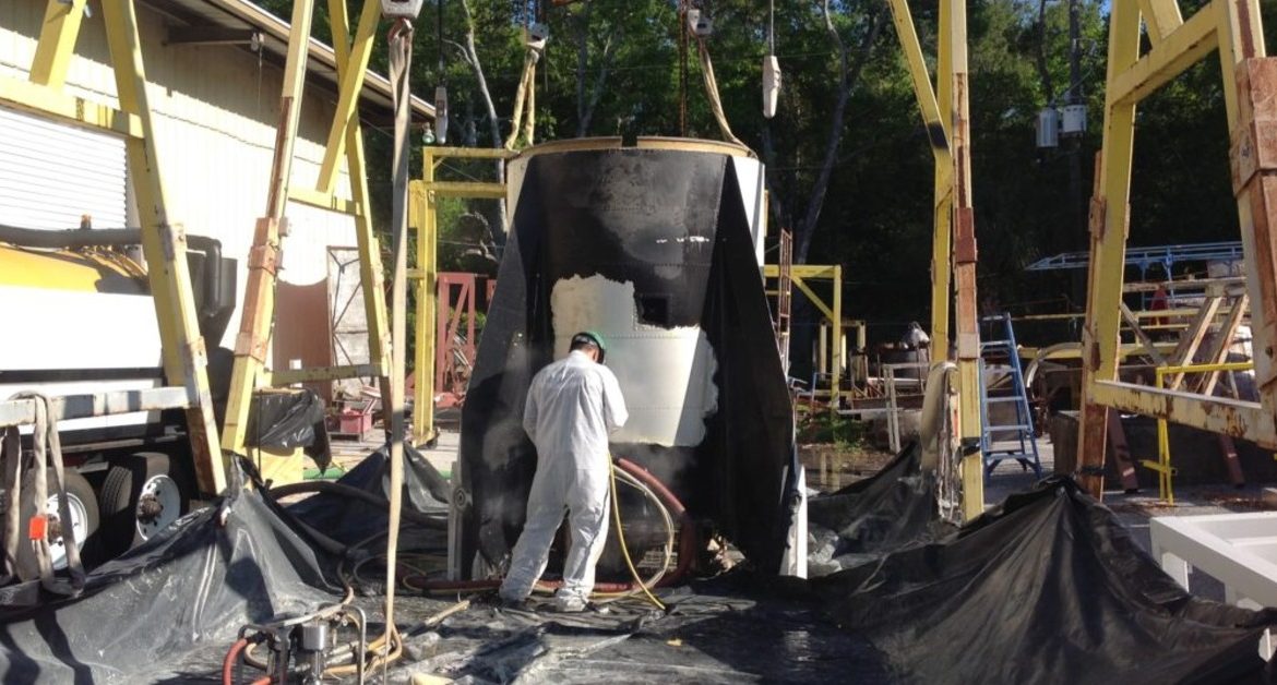 A worker in white coveralls spraying a large black industrial structure outdoors amid equipment and plastic sheeting.