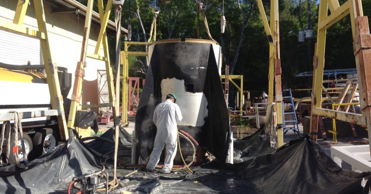 A worker in white coveralls spraying a large black industrial structure outdoors amid equipment and plastic sheeting.