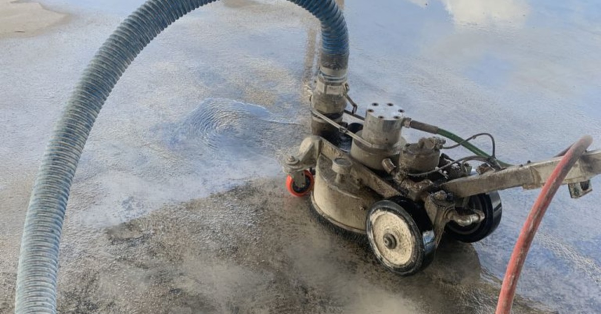 A tool with a brush-like underside and attached blue and red hoses on a wet concrete surface with loose grit nearby.