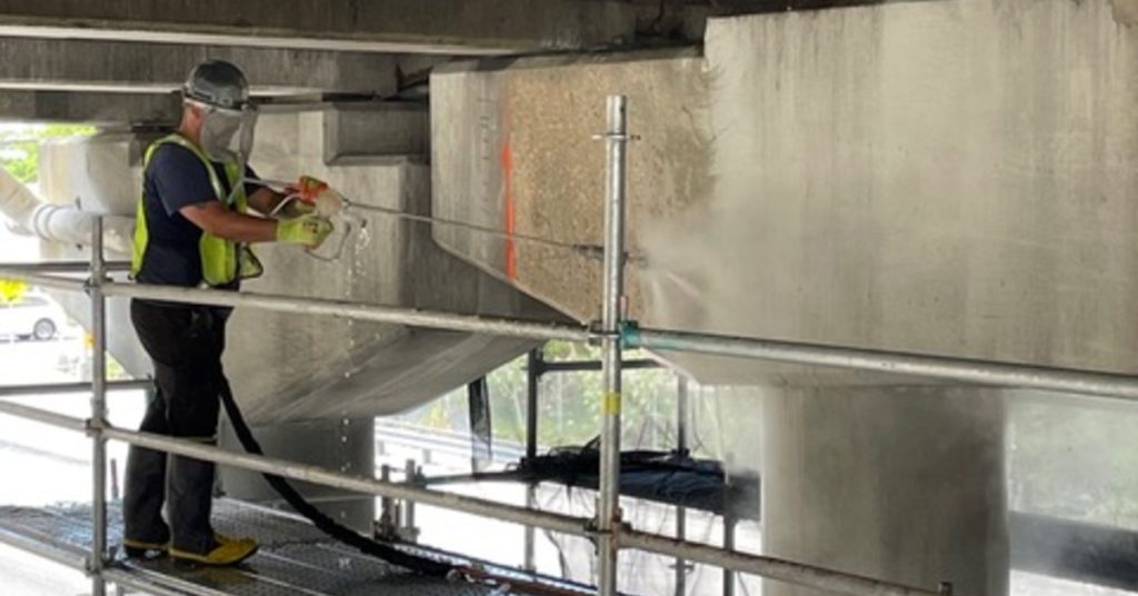 A worker pressure washing concrete beneath an overpass while standing on scaffolding near pillars and beams.