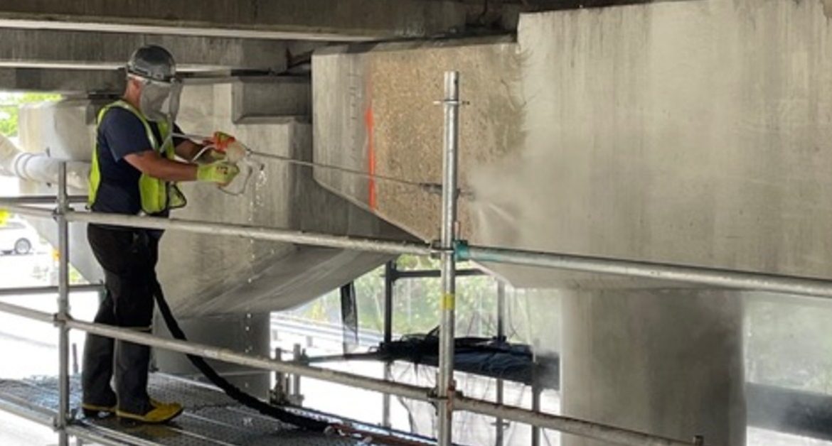A worker pressure washing concrete beneath an overpass while standing on scaffolding near pillars and beams.