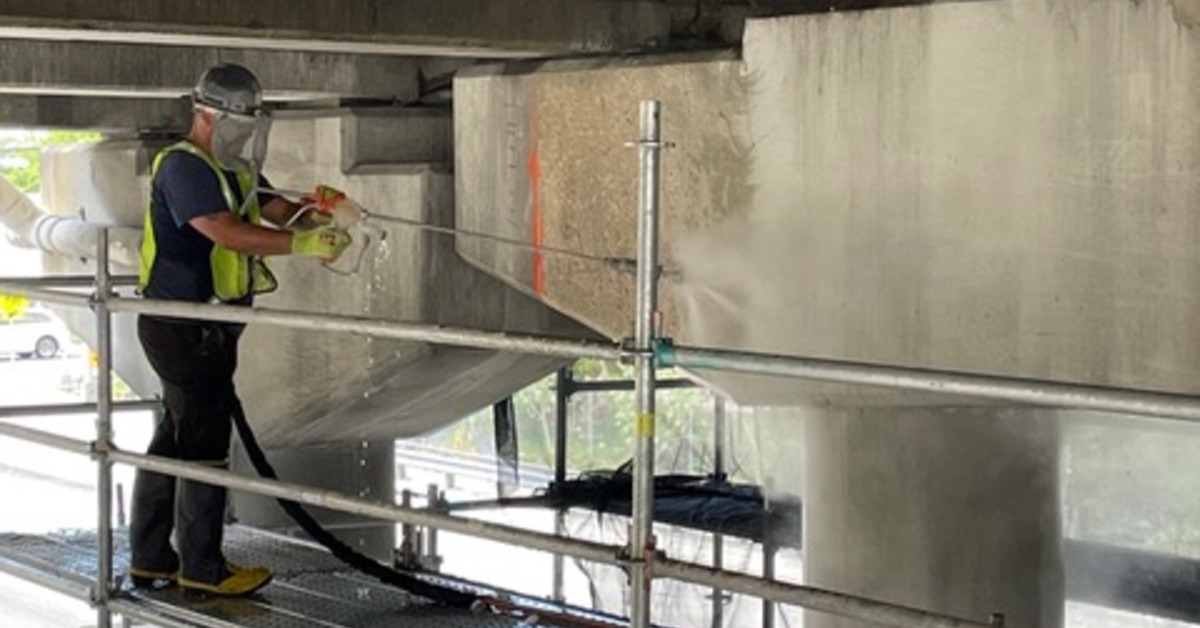 A worker pressure washing concrete beneath an overpass while standing on scaffolding near pillars and beams.