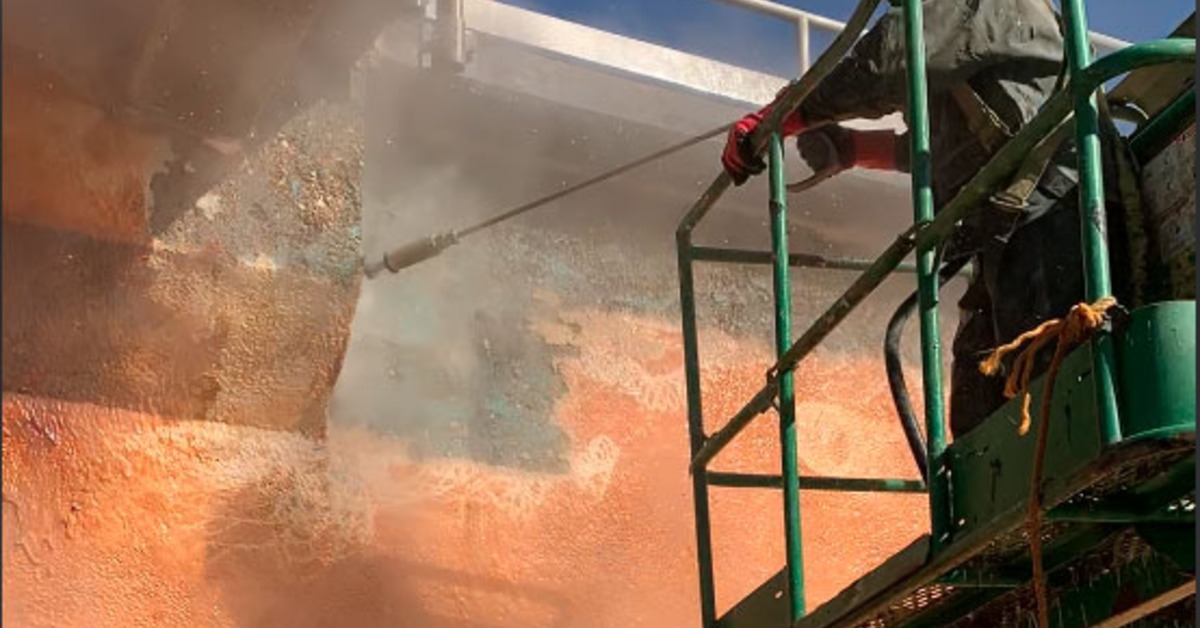 A worker pressure washing a concrete wall from a lift under a bridge with orange surface dust and mist in the air.