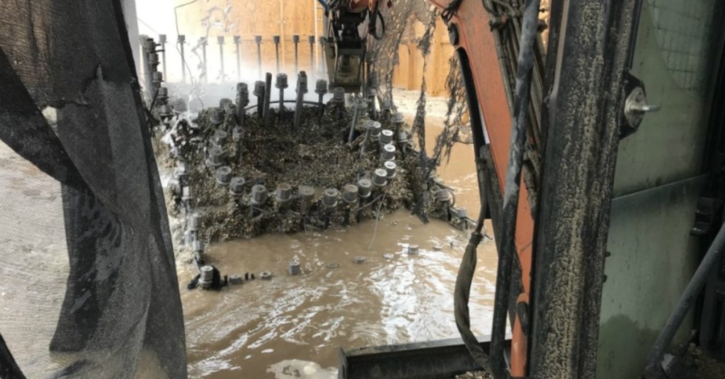 A muddy construction area with machinery, hoses, netting, and standing water around circular metal equipment.