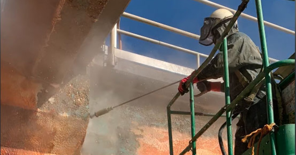A worker wearing protective gear spraying a large rusty structure from a raised platform under a blue sky.