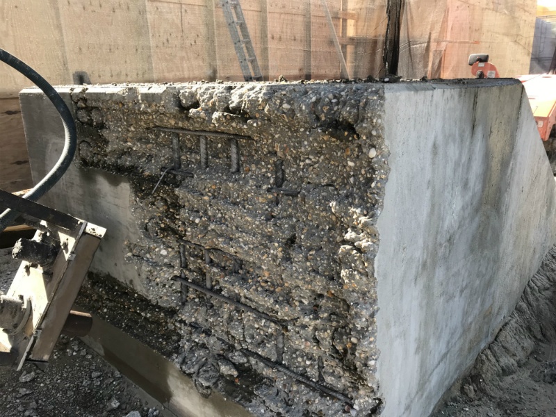 A partially broken concrete block with exposed rebar, rough aggregate, and construction equipment beside it.