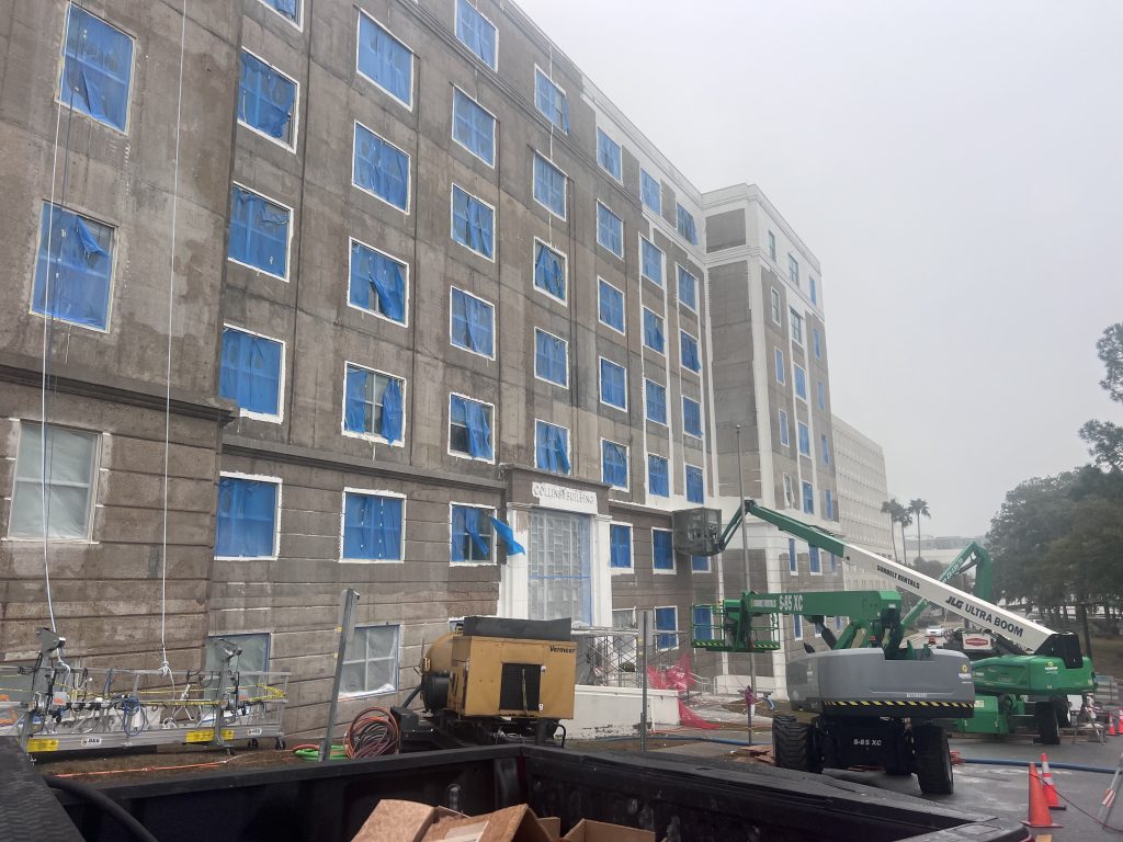 A tall building facade with many blue-covered windows, a green boom lift, construction equipment, and traffic cones.