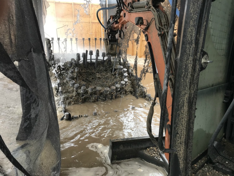 An orange excavator arm working beside muddy water, broken concrete, metal bolts, and black mesh on a jobsite.