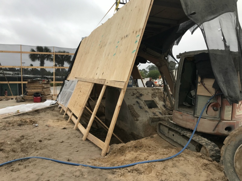 A plywood panel leaning beside an excavator, sand piles, fencing, stacked boards, and a hose at a work area.