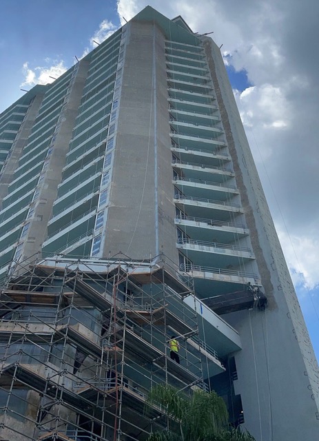 A tall building with glass balconies, exterior scaffolding, suspended platforms, and workers near the lower floors.