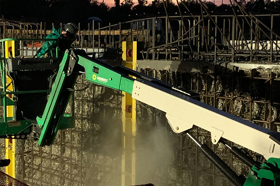 A green boom lift extending toward a concrete wall with exposed rebar, work platforms, mist, and lights at dusk.