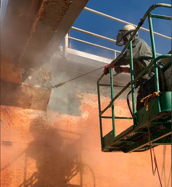 A worker in a green lift is pressure washing a rust-stained metal tank wall. Rising mist surrounds the nozzle.