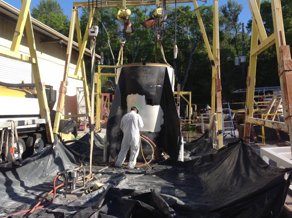 A person in white coveralls is working on a suspended black metal tank section beneath yellow gantry cranes and hoists.